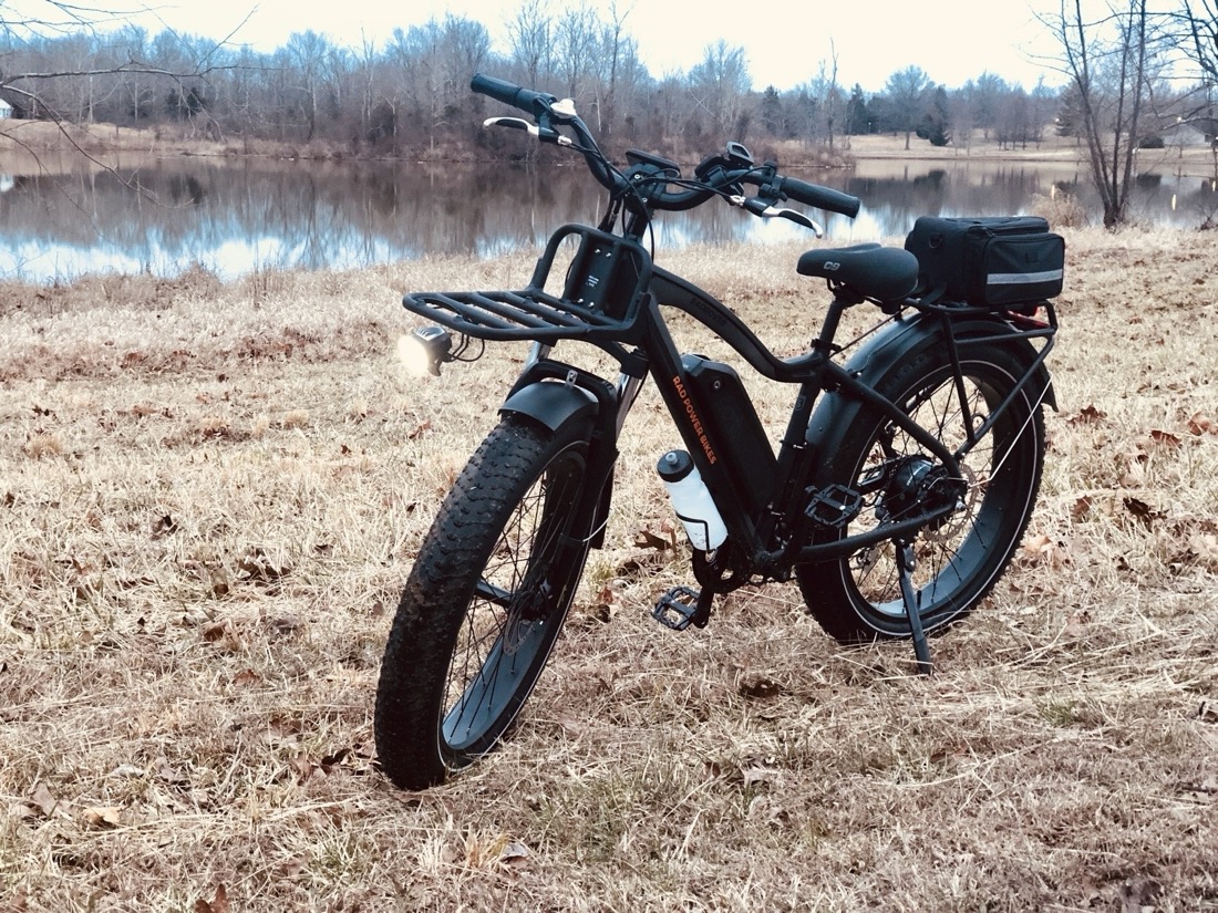 A black fat tire bike on brown winter grass in front of a lake. The bike has a battery pack on the down tube and racks over the front and rear tire. A black pack is attached to the top of the back rack.