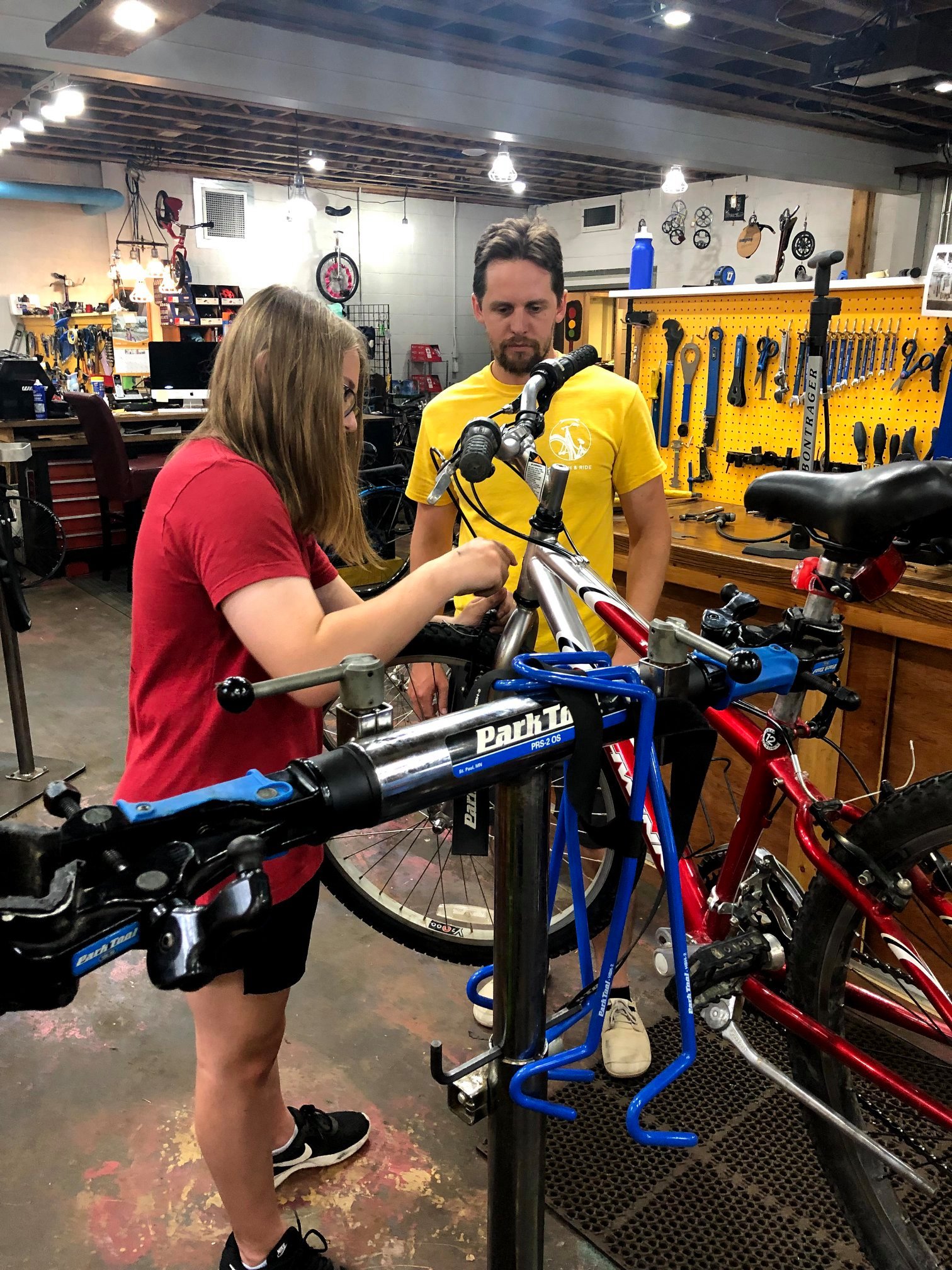 Two people walking and working on a bicycle in a work stand with tools in a nearby workstation