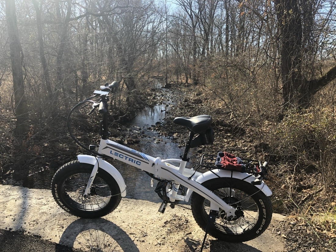 A smaller than average white electric bike with a single rectangular cross tube that contains a battery. The handlebar is held by an unusually tall stem. 