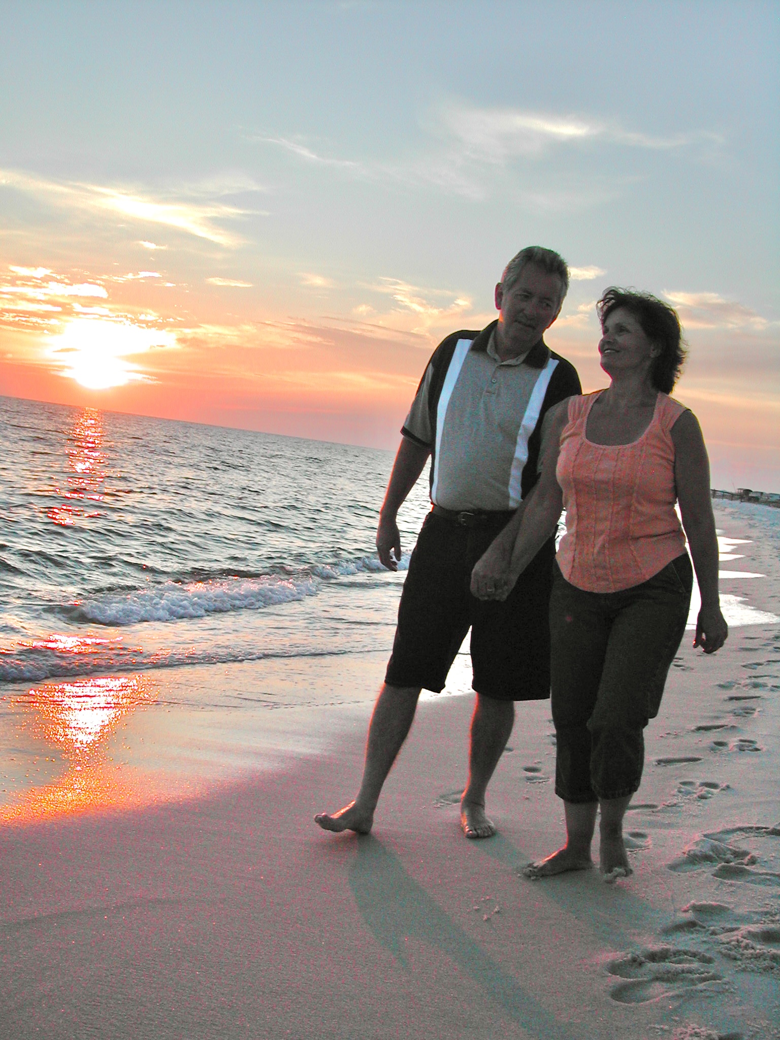 2 people, a man and woman holding hands and walking along the beach at sunset