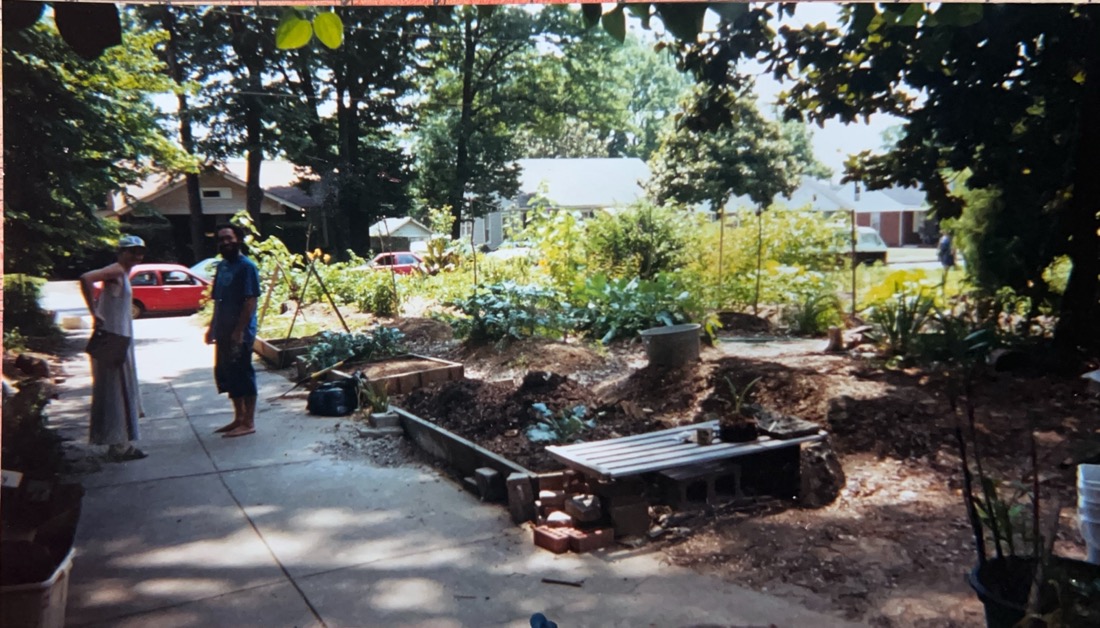 A bearded man and woman wearing a bicycle helmet stand on a driveway next to a large front yard garden.