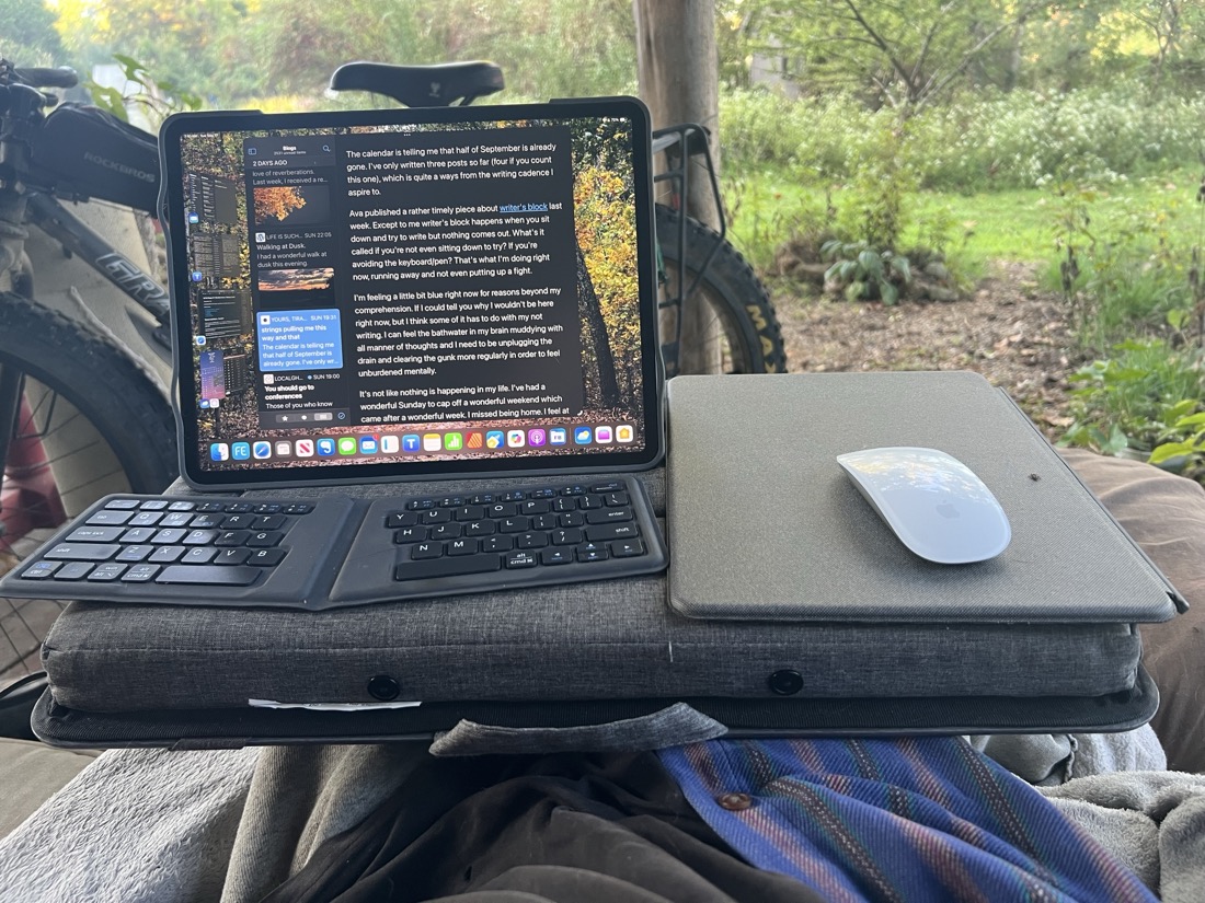 An iPad sitting on a lap desk cushion side up. To right of iPad is a flat gray board with a Magic Mouse. I thin foldable keyboard sits in front of iPad.