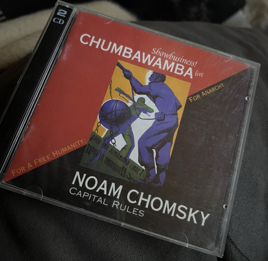 A CD case that is split diagonally. The top half is red with the text: Show business Chumbawamba live. The bottom diagonal is black with the text Noam Chomsky Capital Rules. Along the diagonal line is the text For a free humanity, for anarchy. In the middle of the diagonal split is a box of art depicting two working class men with guns 