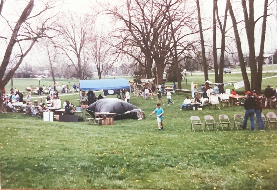 An semicircle arrangement of 40 tables on a large green lawn. At the center appears to be a large black tent which was used to show a film inside 