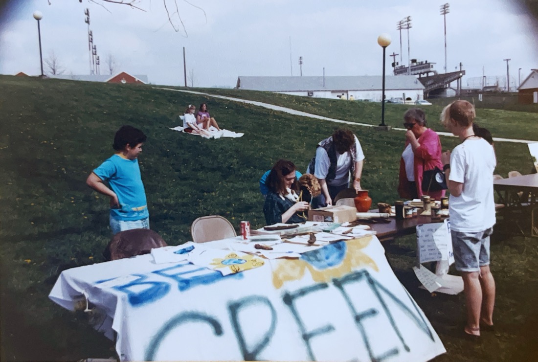 a white sheet banner on a table reads Be Greens. The table has a mix of literature. several people are behind the table 