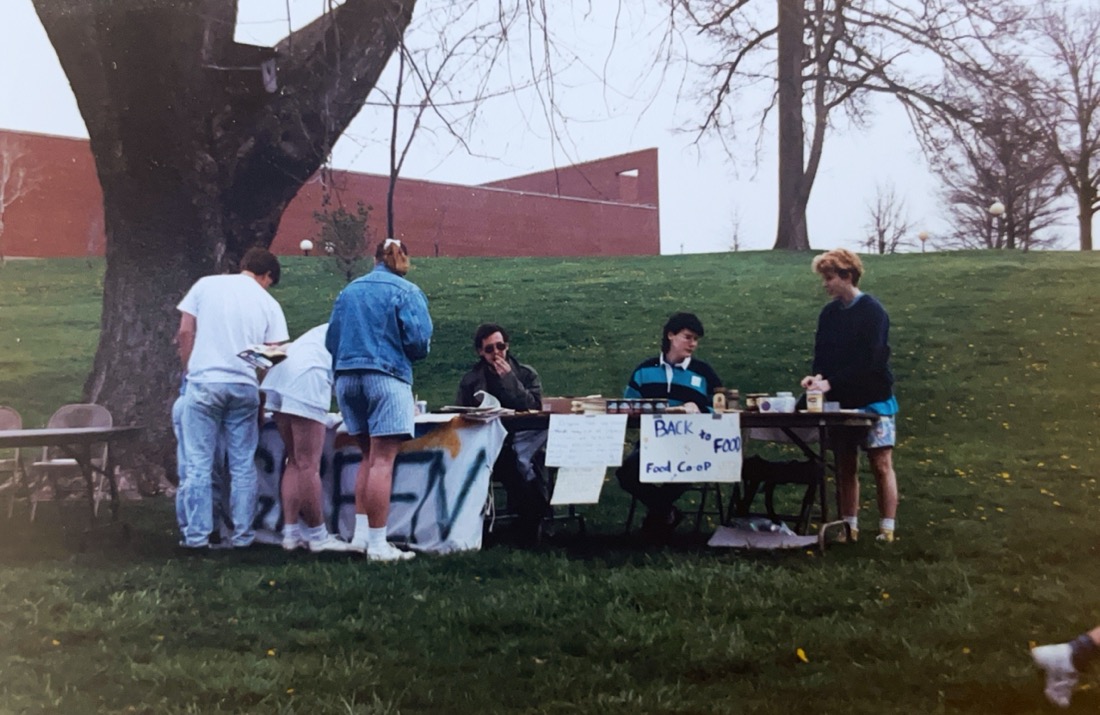 a table sitting on a green lawn. A white banner with the word green is visible draped over the table. People stand in front of and around the table