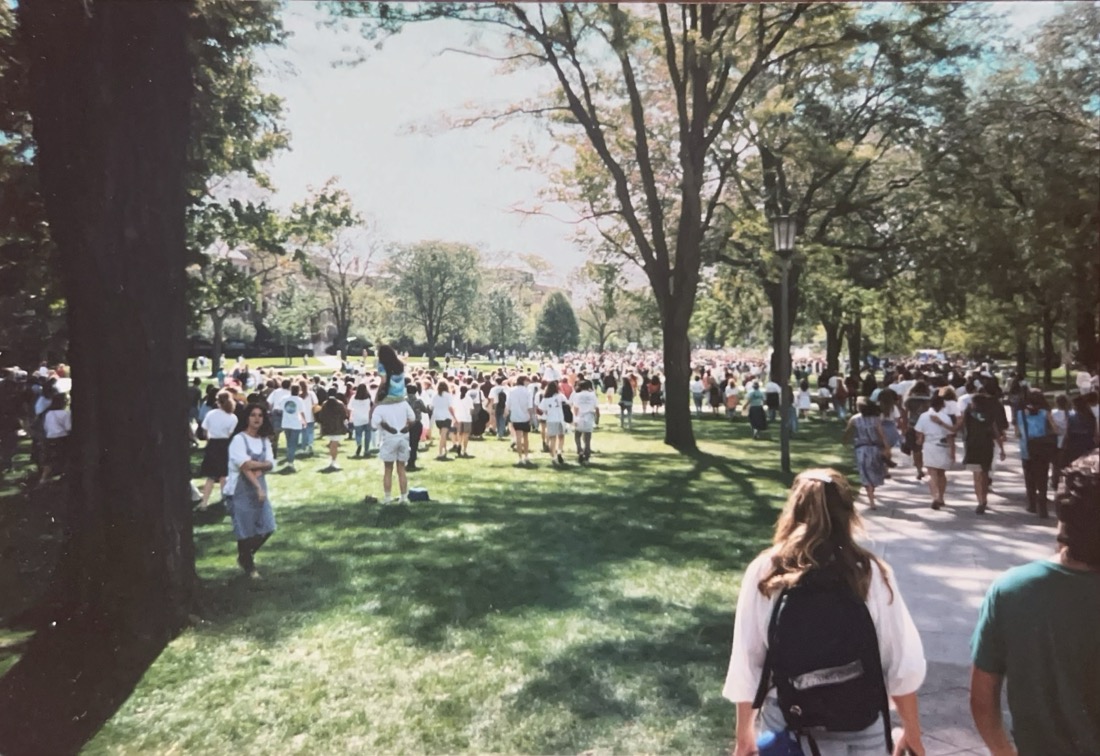 Hundreds of people are seen in a mass walking over grass towards a distant tree line