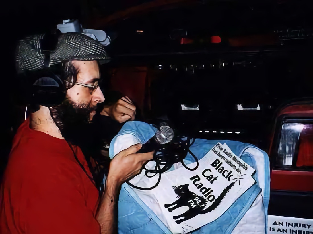 A bearded man in a red shirt and cap wearing headphones and holding a microphone. Behind him is his broadcast equipment is in the trunk of a red car. A flyer in front of him reads Black Cat Radio 94.7FB 