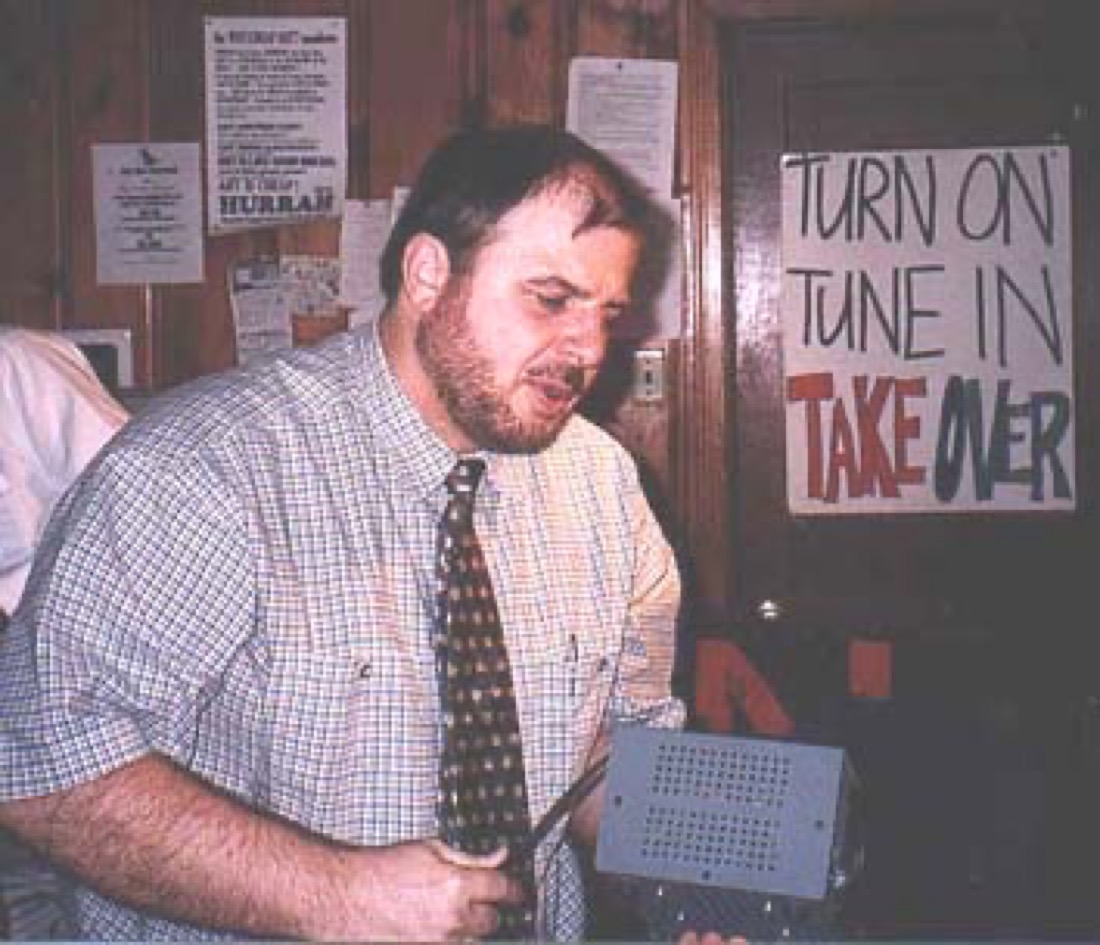 A bearded man in i short sleeve shirt and tie holding radio equipment that does not belong to him which he is in the process of confiscating