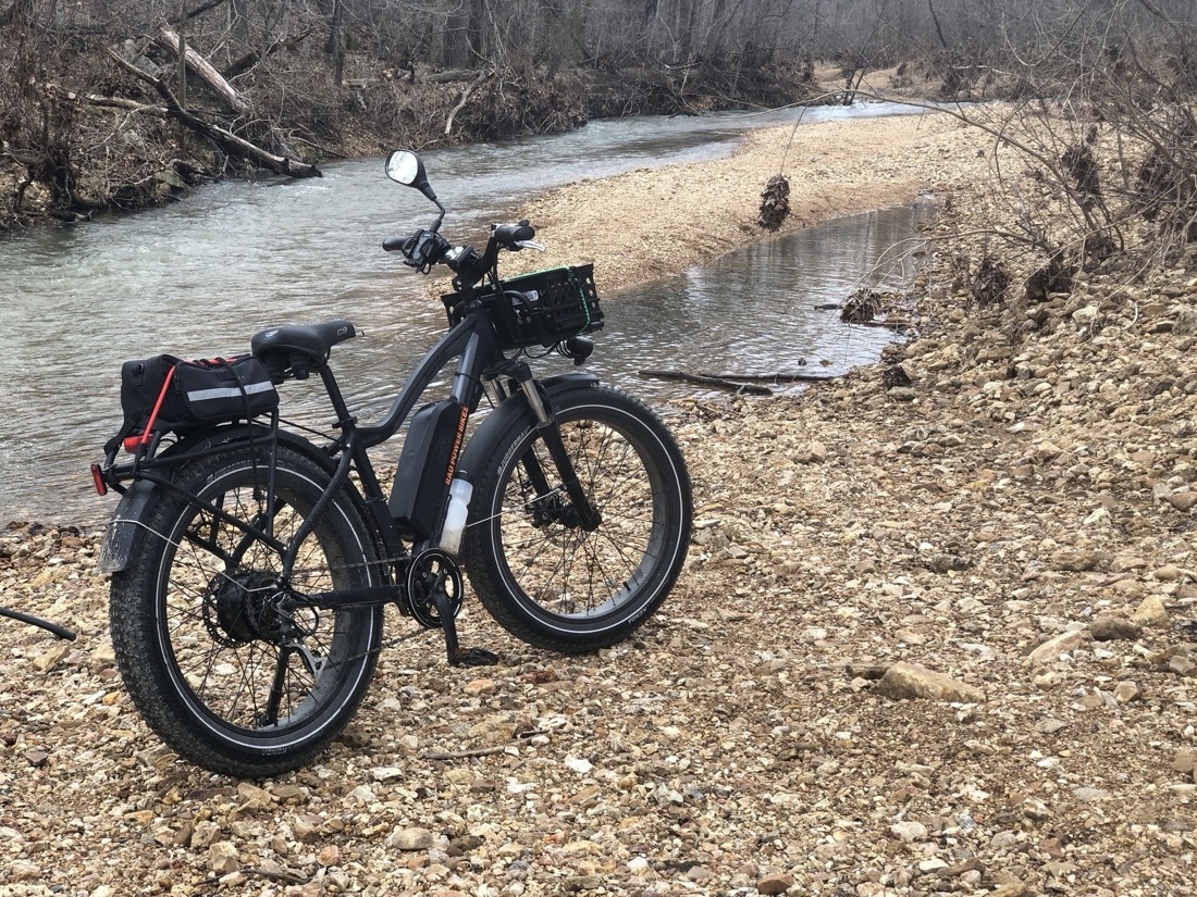 A black fat tire electric bike on a kickstand sits on a sandbar next to a creek