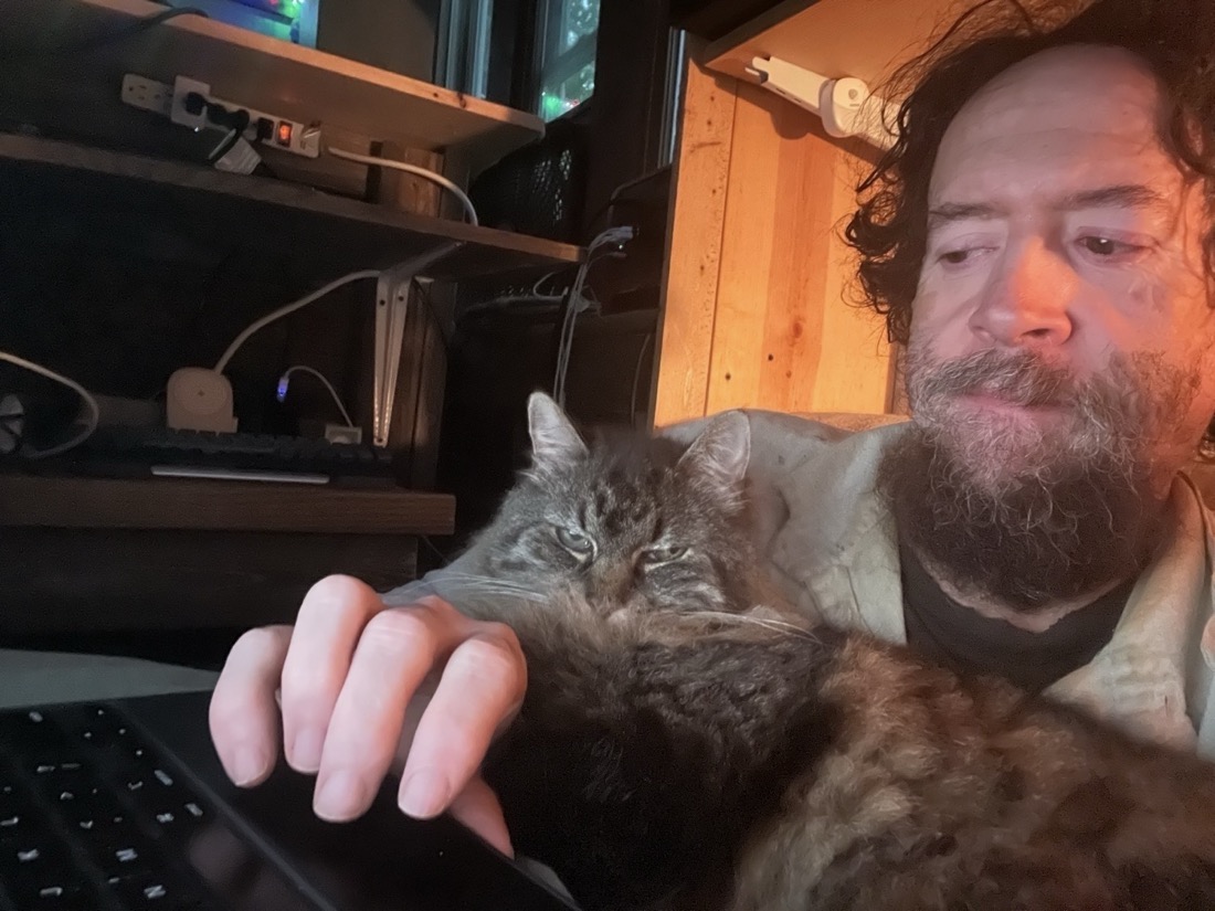 Image taken fairly closeup from an iPad front camera of a brown haired beardy man. He sitting in front of wood shelves and a long haired gray-brown cat lays across his chest and right arm looking at the camera. She is laying across his arm and his hand barely reaches a keyboard at the bottom of the image.