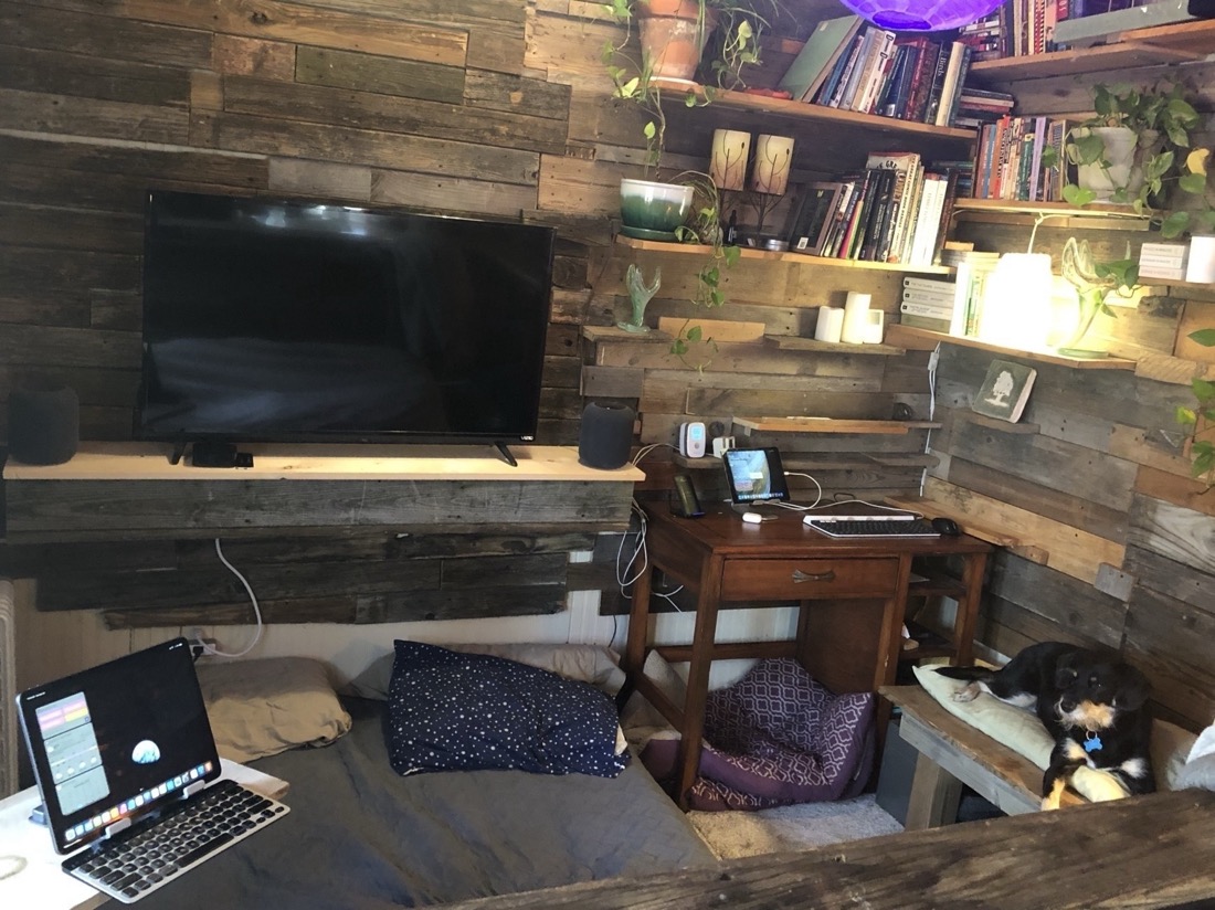 A view of a room. To the right, in the corner is a desk, an iPad and keyboard. To the lower left is a larger iPad Pro in a stand sitting on a white shelf with a keyboard. A dog is sitting on a bench to the right of the photo.