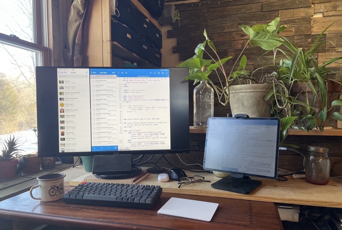 An iPad Pro in stand sitting to the right of a monitor with a keyboard and trackpad sitting in front on a desk. The app Textastic is being used on the monitor to edit a website.