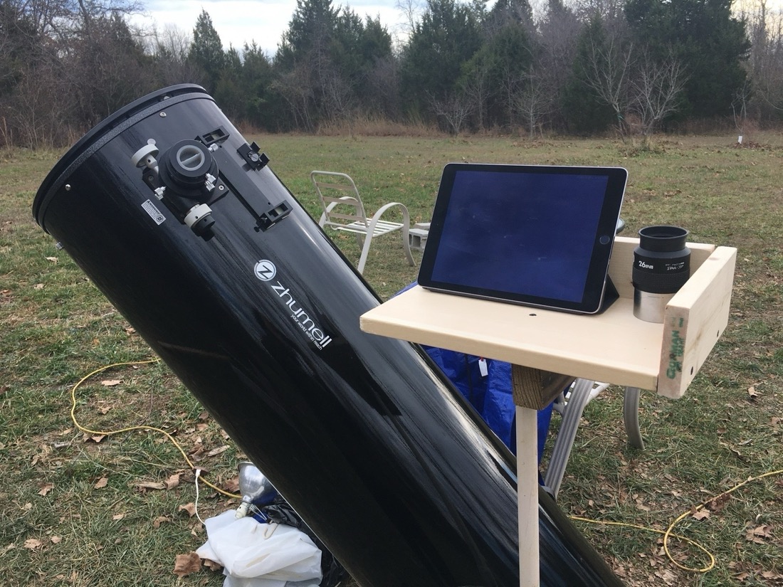 An iPad sits on a wood shelf that is attached to a large dobsonian telescope base which is below the bottom of photo, not visible. The wood plank is held up with a pipe. So that it is at standing level. The telescope is at an angle away from the iPad.