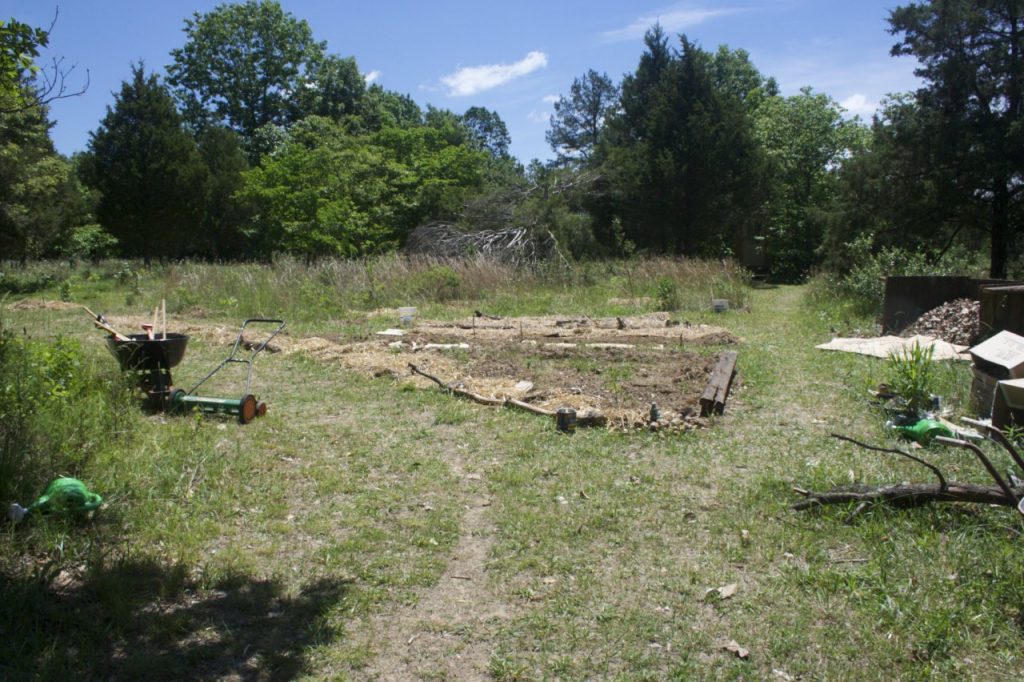 The first days of the garden. Mostly mowed grass with a path to the outhouse on the right side of image. To the right of path is a compost pile. To the left are the beginning garden beds and the the left more cut grass. An unpowered reel style grass mower and wheelbarrow are need the garden. Behind the garden are the tall native grasses of the field.