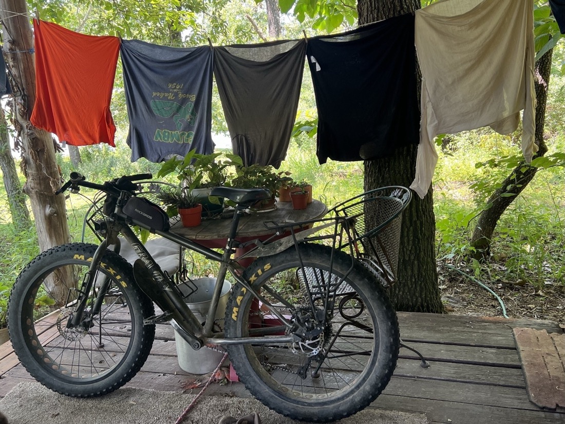 A bike is parked on a porch, leaning against a table. Above it a clothesline with 5 shirts are hanging to dry. Just beyond the porch a woodland of green is the background. 