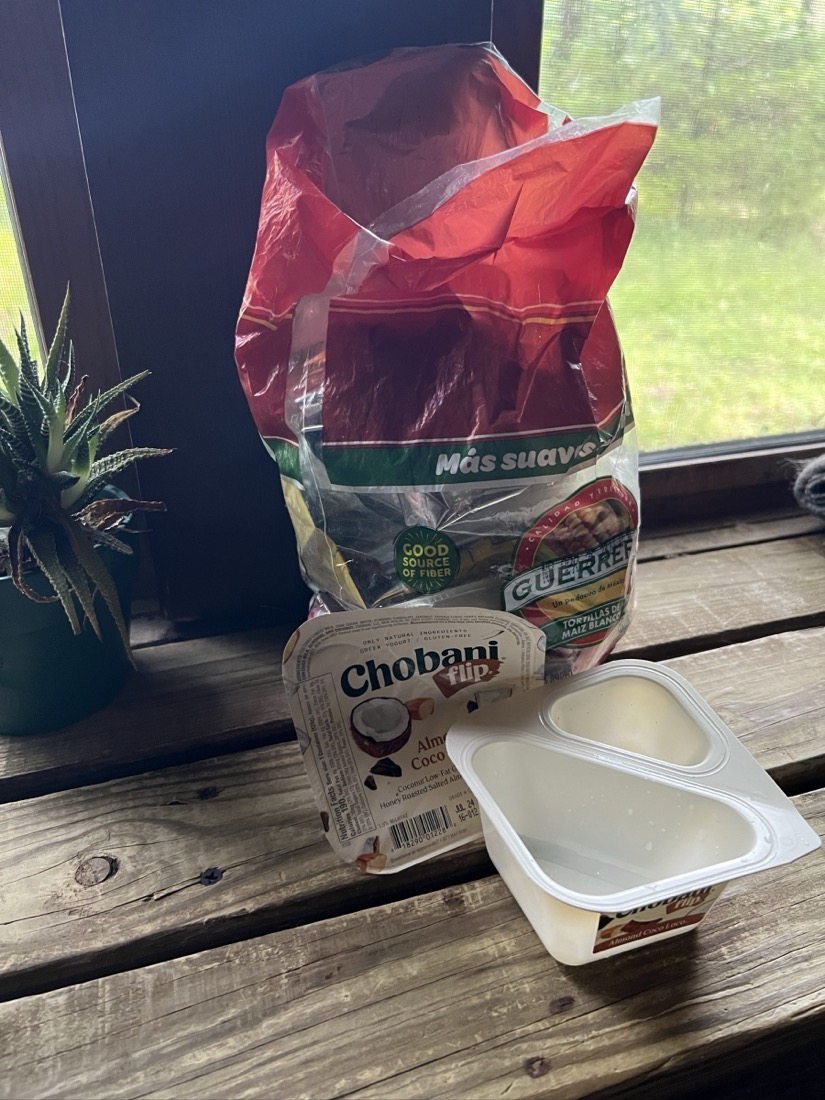 A plastic, single serve yogurt container sits on a wooden shelf in front of a small plastic bag that originally came with corn tortillas. The bag is being repurposed to collect trash and has about three weeks of waste in in it.