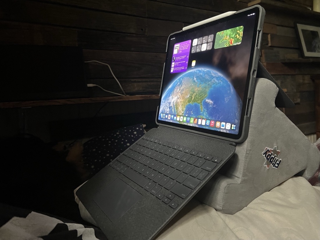 An iPad configured in a Logitech Combo Touch keyboard is propped up on a tablet pillow stand and is being used in a bed