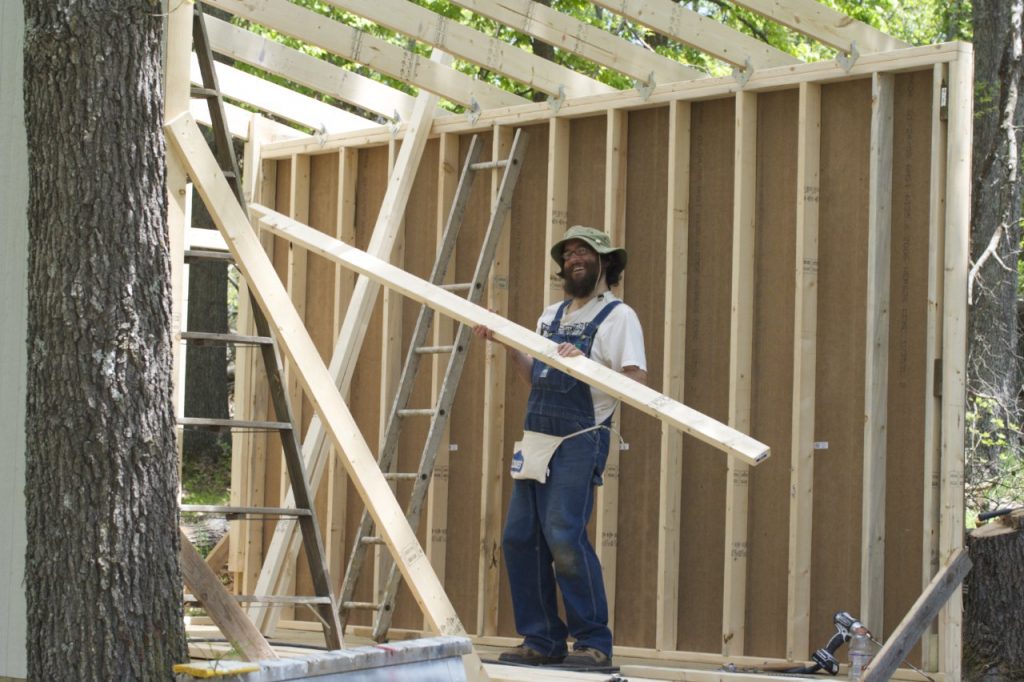 A smiling beardy guy wearing overalls and a green hat carrying 2x4 lumber. One wall of cabin is visible as are roof rafters