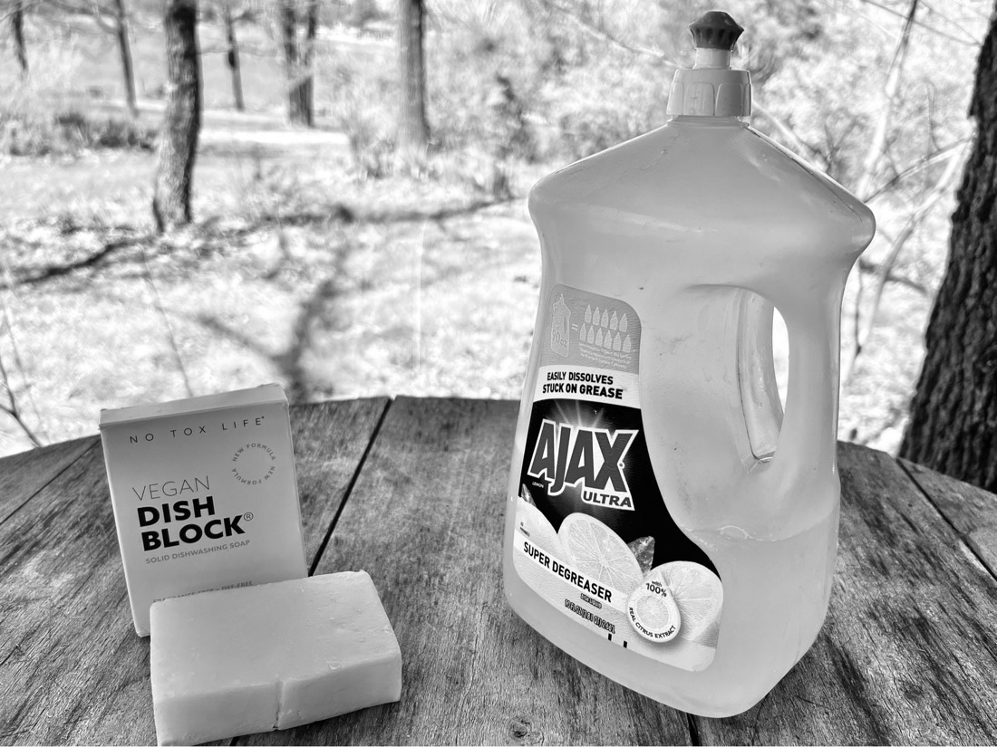 A  white bar of vegan dish soap sits on a wood table next to a plastic bottle of liquid dish soap