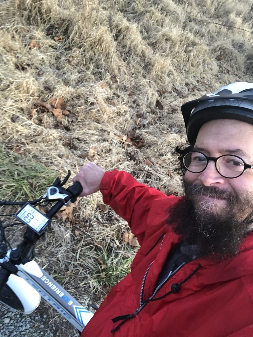 A selfie of a smiling man with a beard and glasses wearing a white cycling helmet and red jacket is leaning against his white bike.