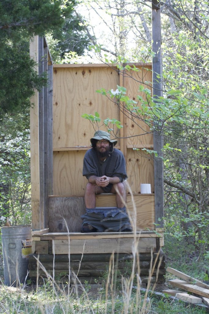 A bearded man sitting in a still under construction outhouse that has no roof or doors