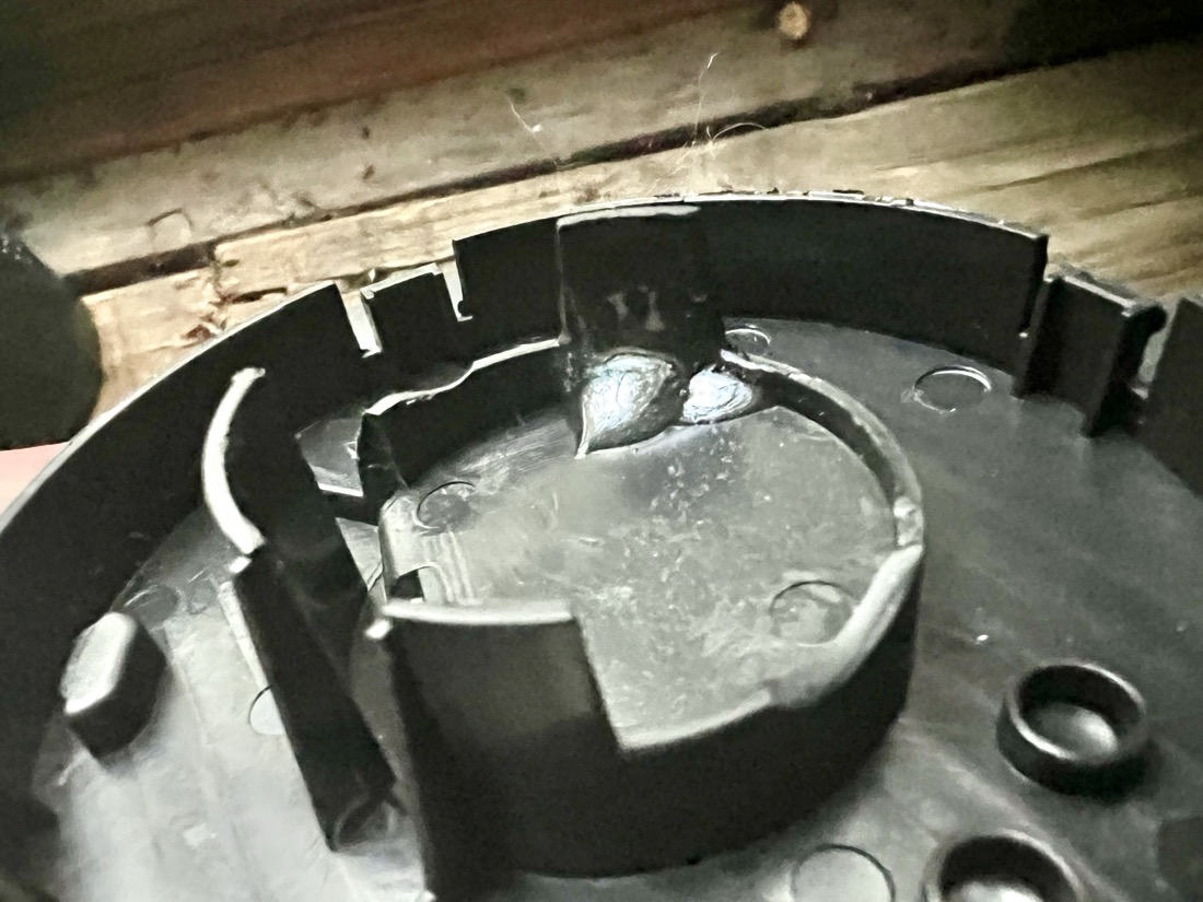 A circular black plastic part about 8 inches in diameter. There are several plastic elements such as a circular ridge of black plastic with tabs in the center. There appears to be a large drop of water along the edge of the inner circle of plastic which is, in actuality, dried glue