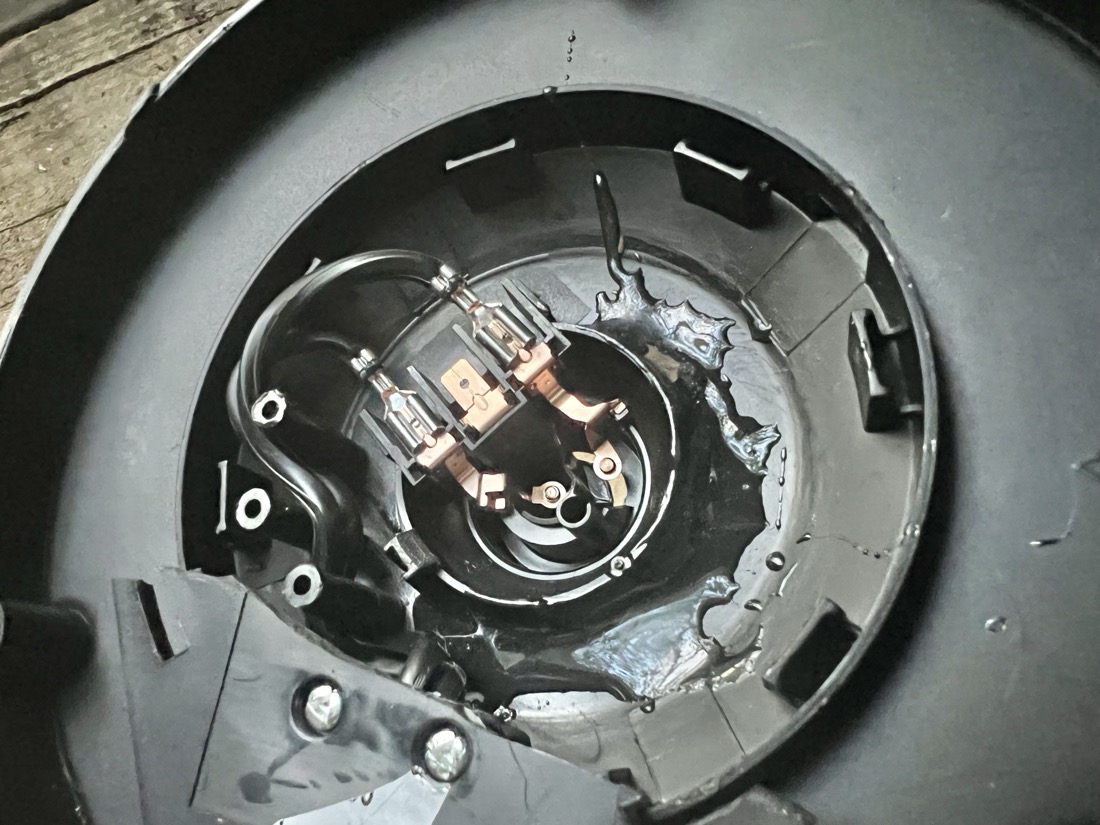 A circular black plastic part about 8 inches in diameter. There are several plastic elements such as a circular ridge of black plastic with tabs in the center. There is a black wire coiled into the center which splits into two and is connected to two copper colored metal clips. There is clear glue all around the inner plastic ridge where the two broken pieces have been glued together 