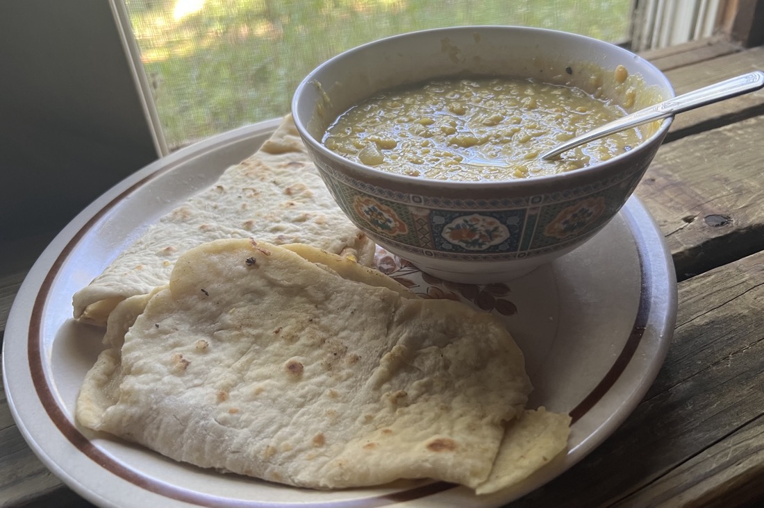 a white plate with a bowl of yellow soup and two pieces of folded flat, naan bread