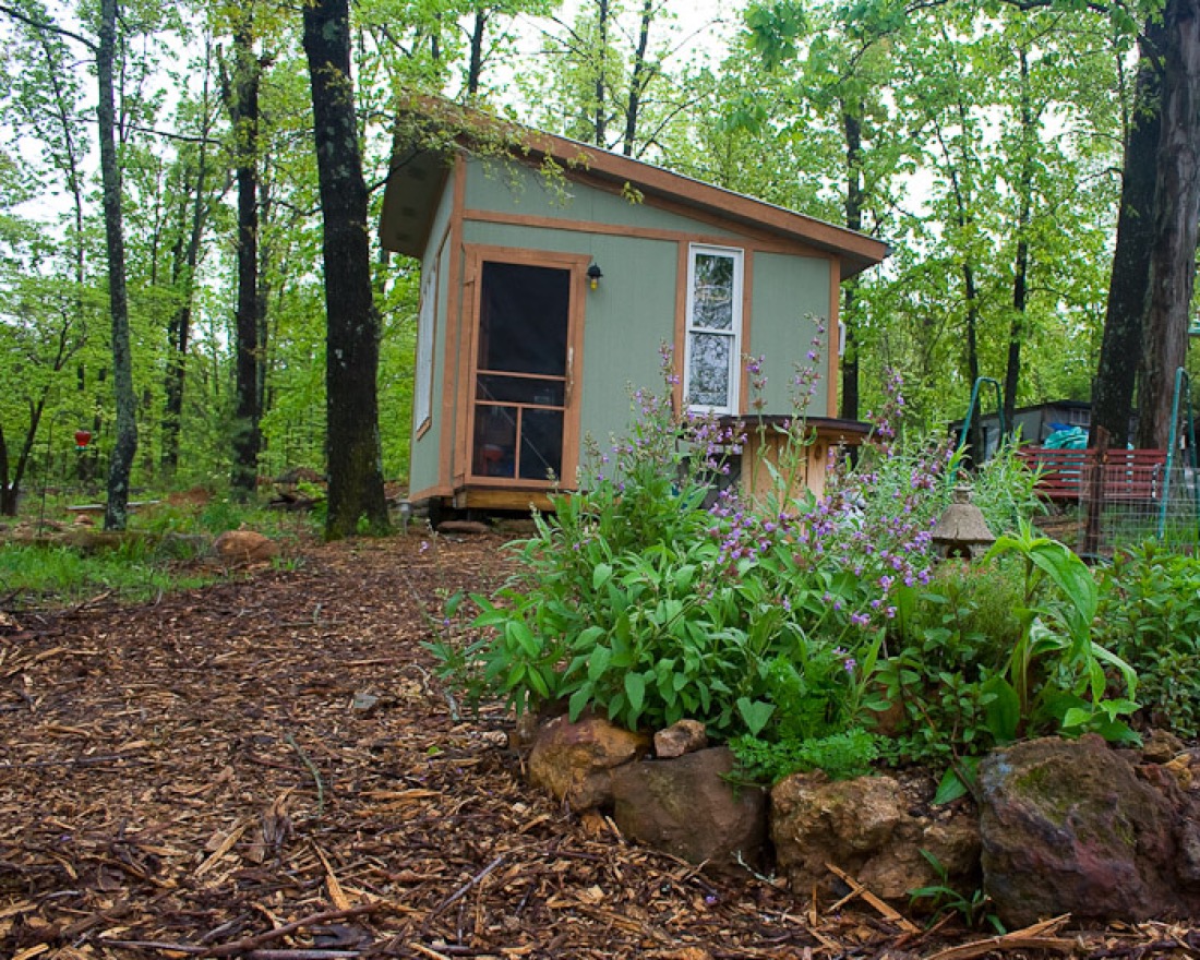 In the foreground a mount of green herbs growing within a border of cantaloupe sized rocks that form a border. In the background is a green cabin.. The scene is set in a shady wooded area