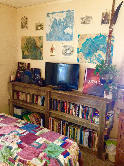 Rustic wood shelves with a small flat screen tv on top, books fill the shelves and vintage drawings and maps are on the wall  