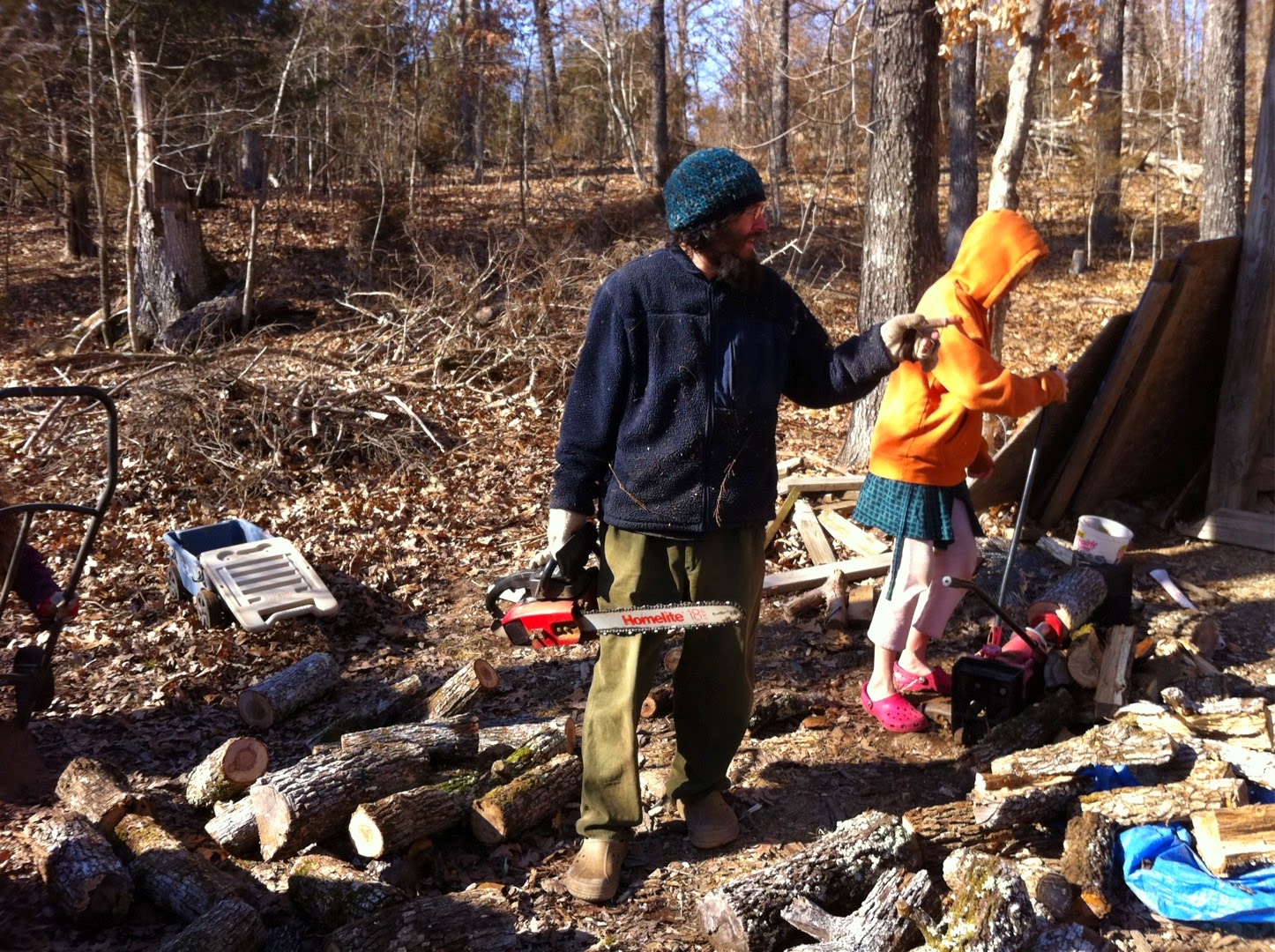 a man in dark blue jacket carries a red chainsaw and is working in front of a woodshed. Various logs surround him. A girl wearing an orange hoodie is behind him using a wood splitter.