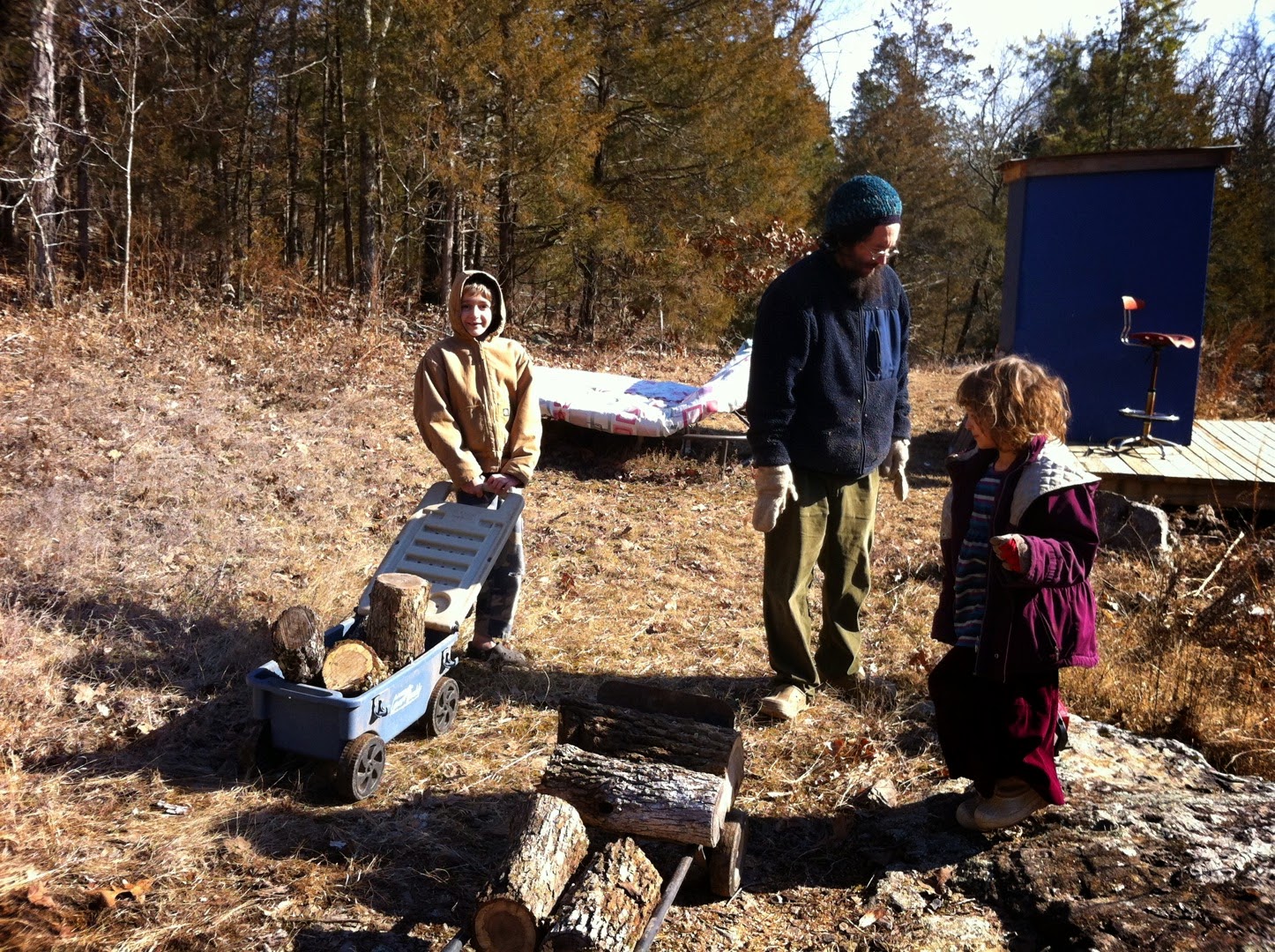 a man in dark blue jacket is looking at something on the ground. Around him are various cut logs. A young boy in a brown coat smiles at the camera as he tugs a wagon with logs. A young girl with curly brown hair in a vibrant purple pink jacket is looking at the wagon.