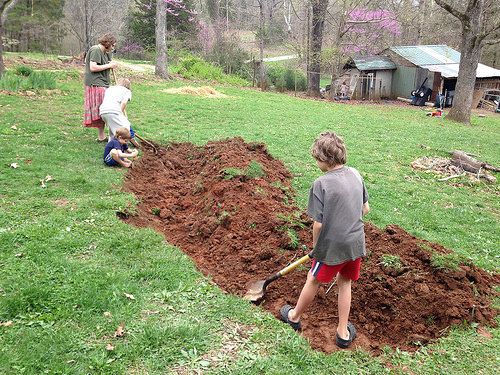 A new water collecting swale on a grassy lawn being dug by kids. It looks like a long ditch in red clay with the dirt dug out piled in a bed along the back side.