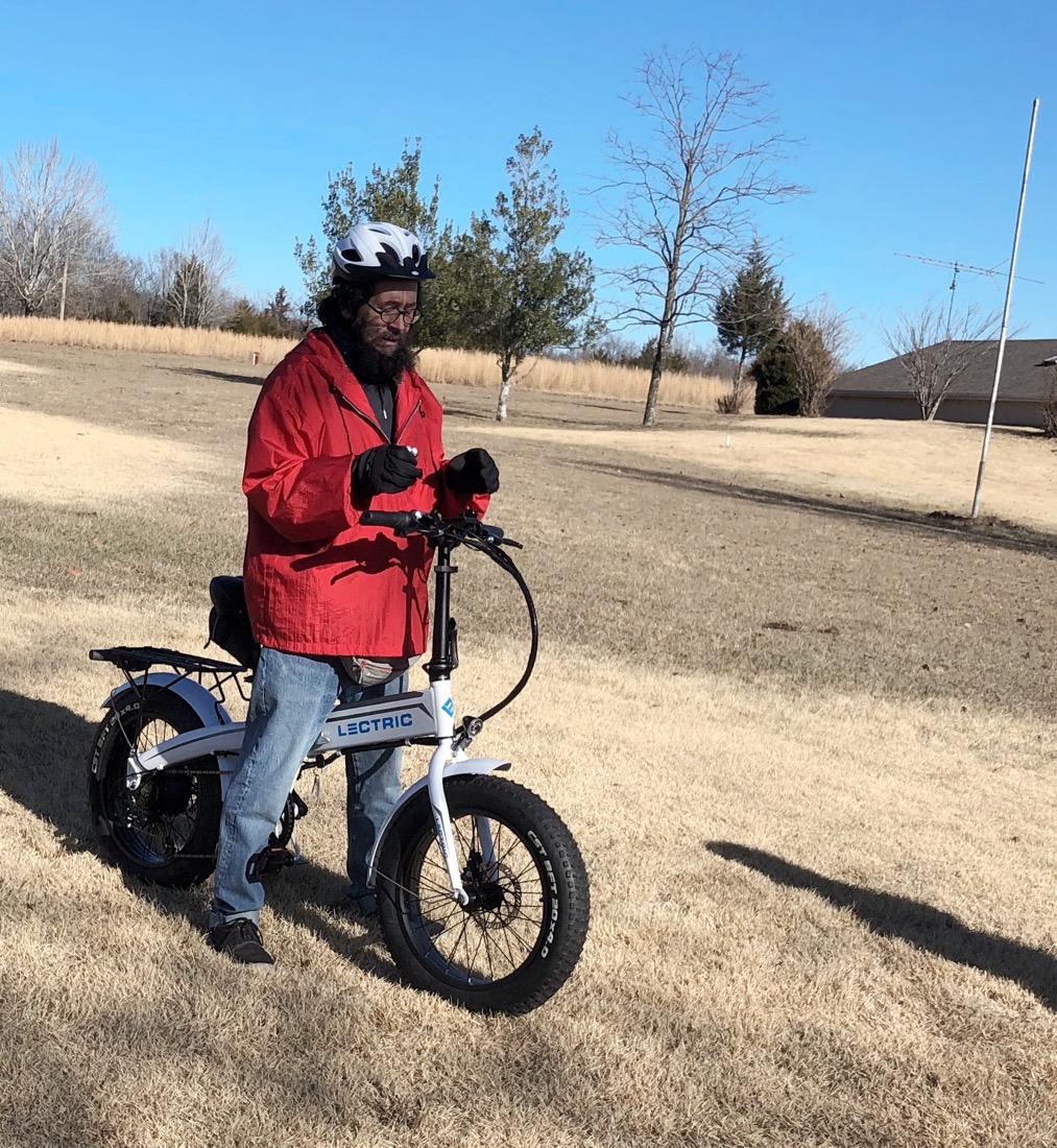 A beardy guy wearing a bicycle helmet and red jacket stradles a white bike