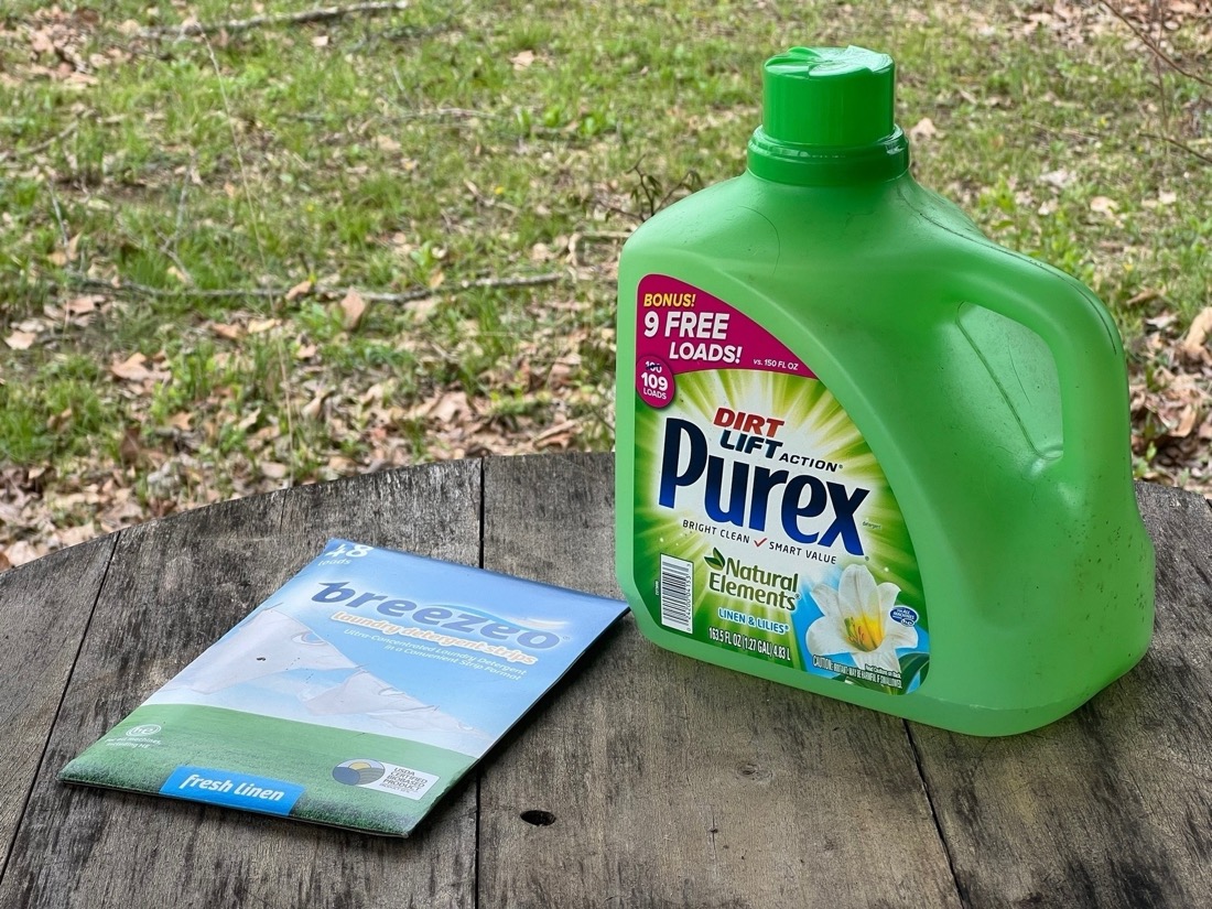 a paper board container of laundry detergent strips is in slim package sitting on the table next to a green plastic jug of liquid laundry detergent