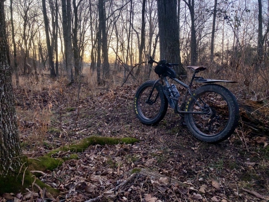 A fat tire bike with multiple water bottle cages, handlebar attached bags and fenders leans against a tree in the winter woods.