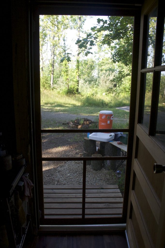 My outdoor sink photographed from inside the cabin. An orange plastic water cooler rests on a small white sink, the whole arrangement is stacked on several large logs that bring the sink to waist height 