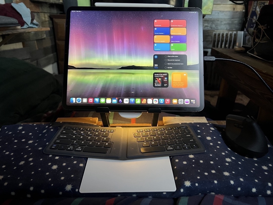 An iPad attached To a stand which is attached to a wooden lap desk. The wooden lap desk has a fabric covering over the wood. A keyboard and trackpad are below the iPad.