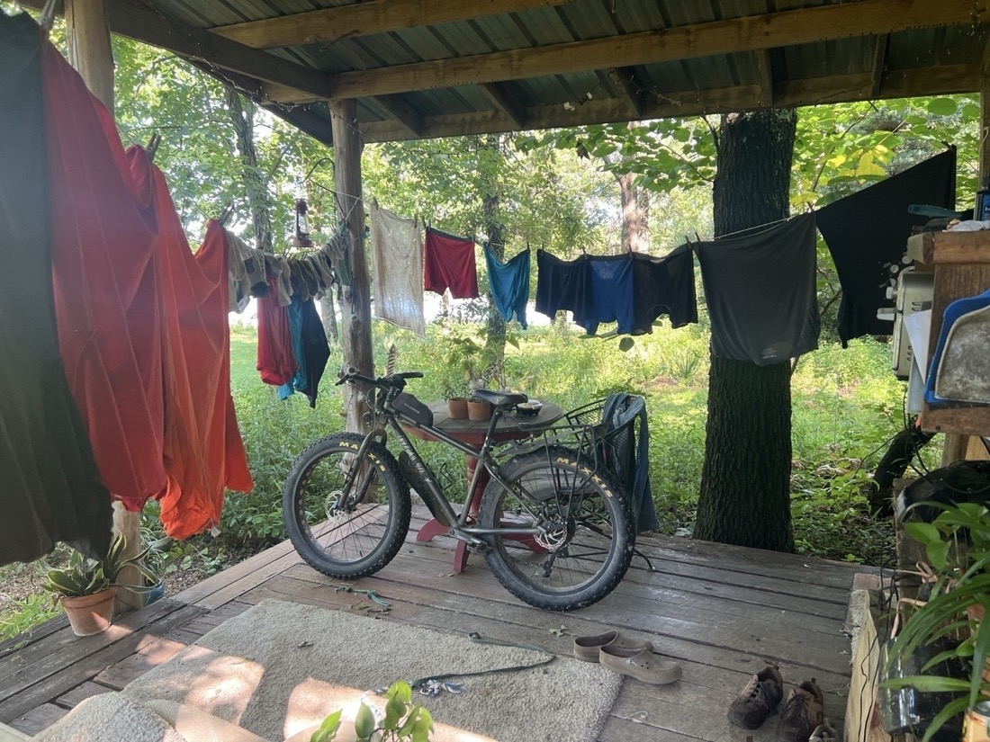 Two laundry lines strung across a wood porch deck with a variety of colorful clothing hanging to dry. Also on the porch, a fat-tire bike leans against a table. There are several shoes visible on the deck and a woodland in the background