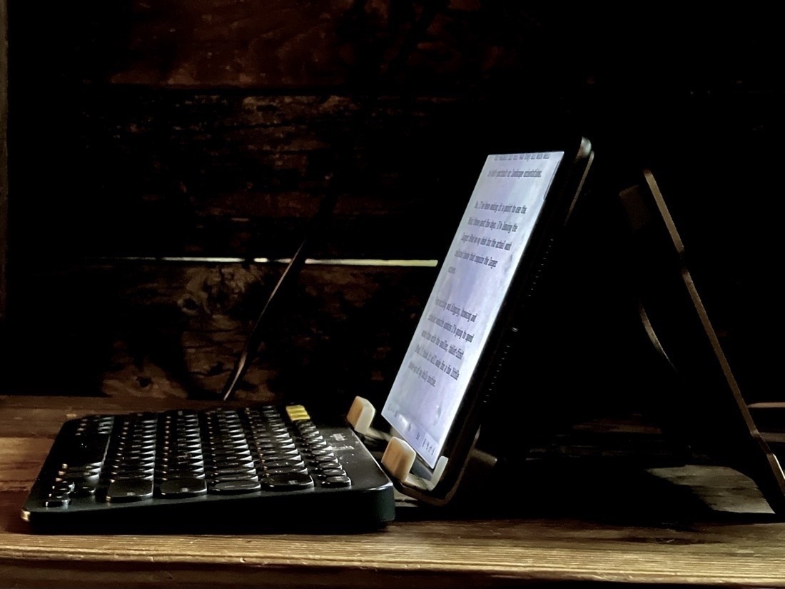 An iPad Mini sits in a stand on a wood shelf. The iPad is facing left, a black keyboard is in front of it