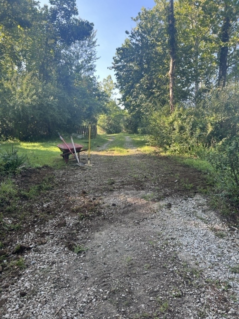 A gravel road looks a bit messy as dirt has been pulled across it to level it. A wheel barrow and tools are off to side of road